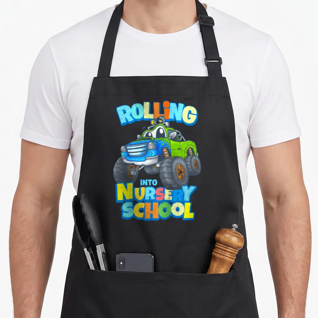 Nursery truck apron - Cartoon monster truck with rolling into nursery school text in blue, yellow, and green — shown on a black cotton canvas apron against a white background
