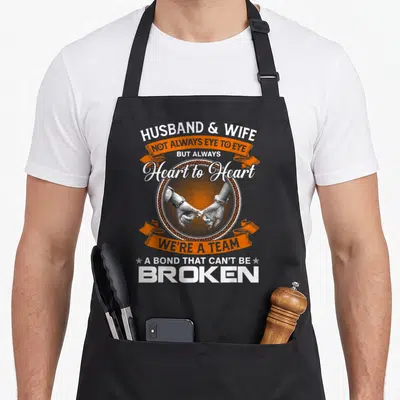 Husband wife apron - Pinky promise hands in white circle with orange ribbons and couple bond quote — shown on a black cotton canvas apron against a white background