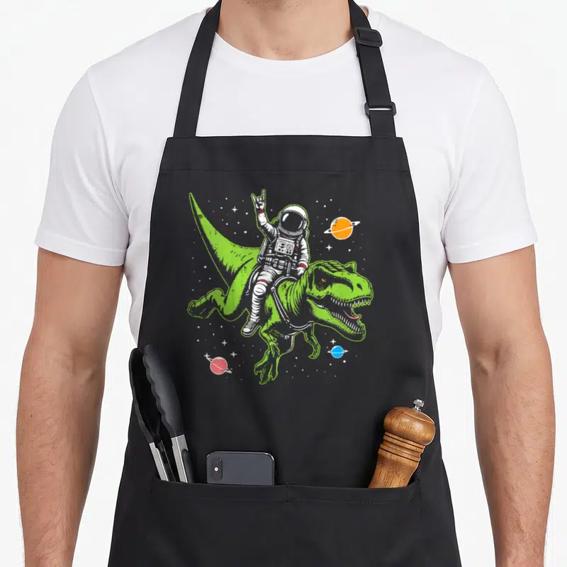 Dino astronaut space apron - Cartoon astronaut riding green T-Rex with three planets and stars — shown on a black cotton canvas apron against a white background