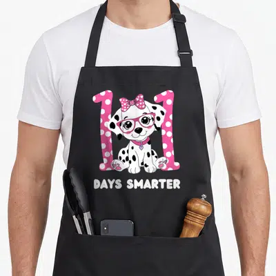 Friendly dog apron - Dalmatian puppy between pink number 1s with DAYS SMARTER text — shown on a black cotton canvas apron against a white background