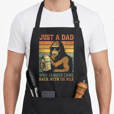Bigfoot milk aprons - Retro sasquatch in sunglasses holding milk carton against orange and yellow stripes — shown on a black cotton canvas apron against a white background