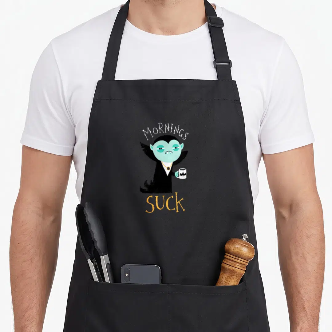 Morning vampire apron - Cartoon vampire holding bat mug with Mornings Suck text in white and orange — shown on a black cotton canvas apron against a white background
