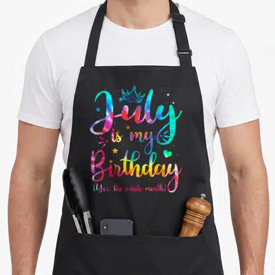July whole birthday apron - Tie-dye cursive text 'July is my Birthday' with crown, stars, heart in pink blue yellow — shown on a black cotton canvas apron against a white background