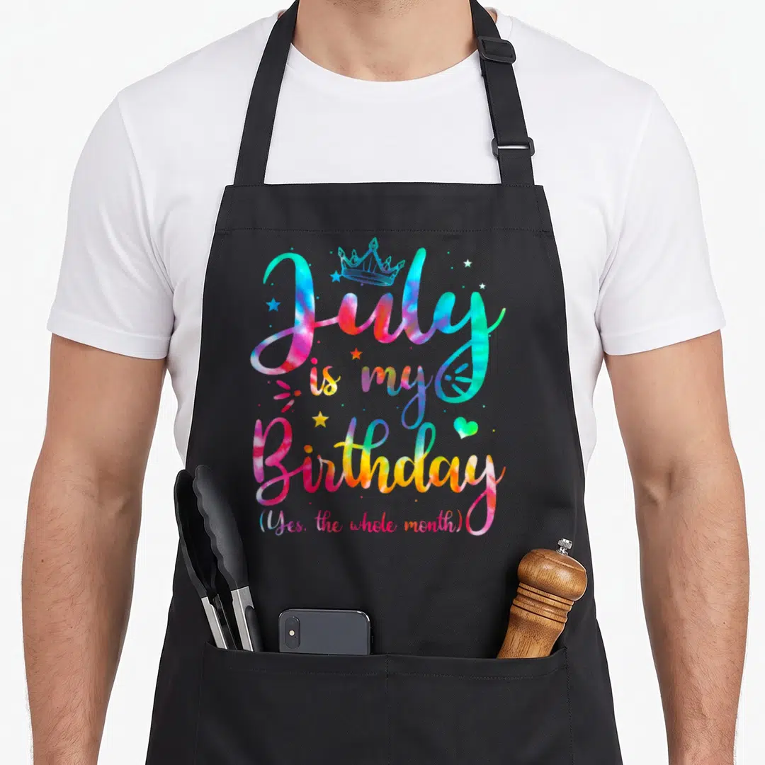 July whole birthday apron - Tie-dye cursive text 'July is my Birthday' with crown, stars, heart in pink blue yellow — shown on a black cotton canvas apron against a white background