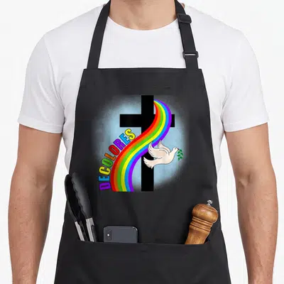 Decolore cross apron - Black cross with rainbow arc, DECOLORES text, white dove holding olive branch in cartoon style — shown on a black cotton canvas apron against a white background