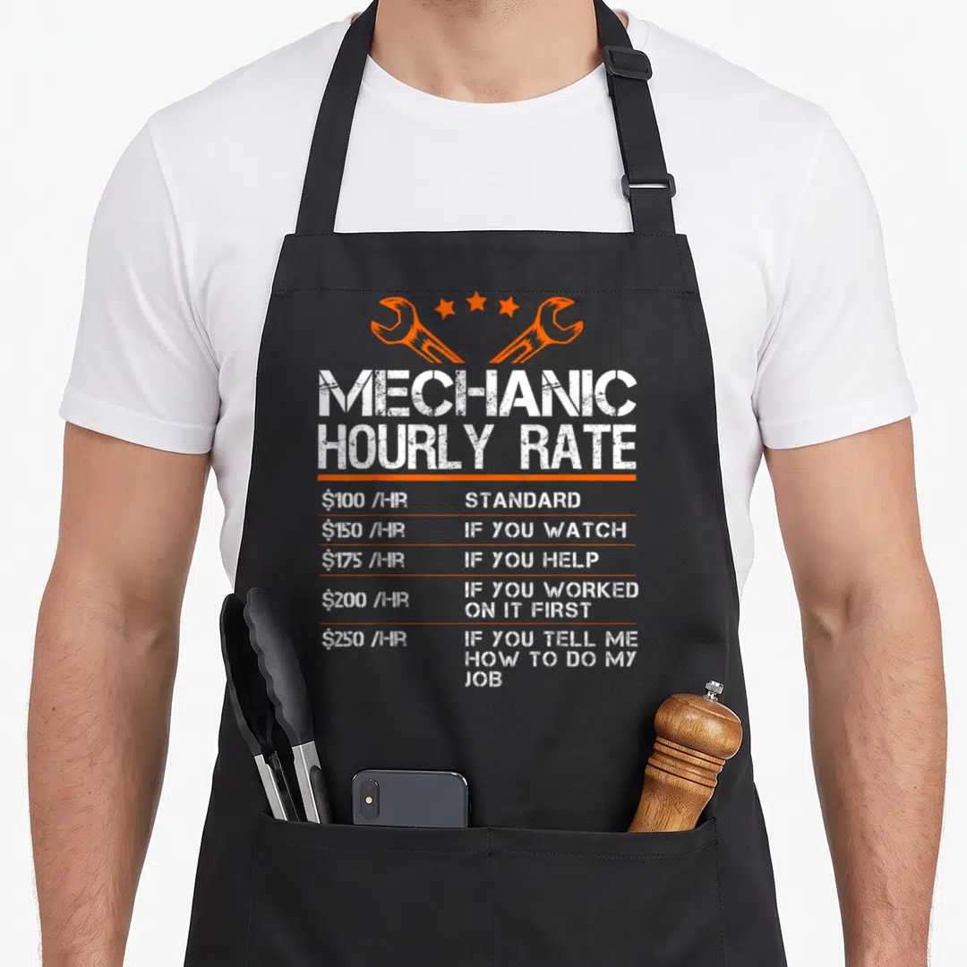 Hourly auto mechanic apron - White distressed text listing mechanic hourly rates under two orange wrenches and three — shown on a black cotton canvas apron against a white background