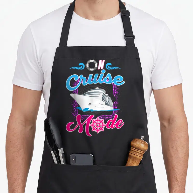 Cruise sightseeing apron - ON Cruise Mode text with ship, lifebuoy, wheel, purple glitter, pink cyan white — shown on a black cotton canvas apron against a white background