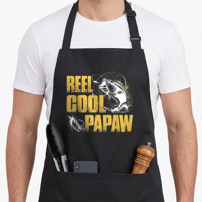 Reel papaw fishing apron - Distressed yellow REEL COOL PAPAW text with white jumping fish and lures — shown on a black cotton canvas apron against a white background