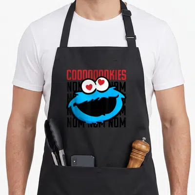 Coooooooky cookie monster apron - Blue cartoon face with red heart eyes over four rows of black text and red header — shown on a black cotton canvas apron against a white background