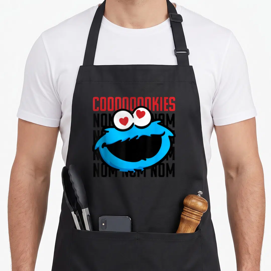 Coooooooky cookie monster apron - Blue cartoon face with red heart eyes over four rows of black text and red header — shown on a black cotton canvas apron against a white background