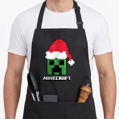 Creeper santa aprons - Pixelated green creeper head in red santa hat above white MINECRAFT text on black background — shown on a black cotton canvas apron against a white background