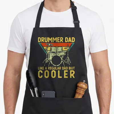 Drummer music dad apron - Retro drum kit illustration with yellow text reading Drummer Dad Like A Regular Dad — shown on a black cotton canvas apron against a white background