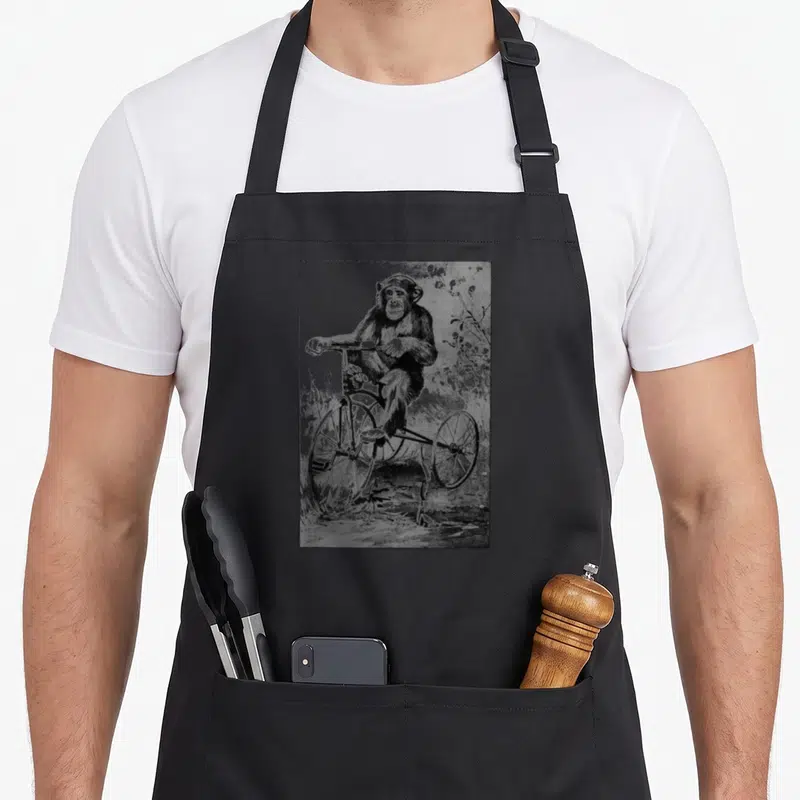 Monkey chimpanzee apron - Realistic chimpanzee sitting on a tricycle in grey and black sketch style — shown on a black cotton canvas apron against a white background
