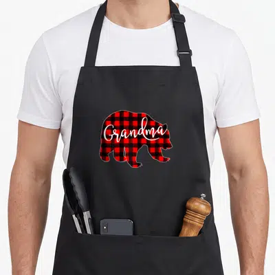 Grandma buffalo plaid christmas apron - Bear silhouette filled with red and black plaid pattern and white Grandma — shown on a black cotton canvas apron against a white background