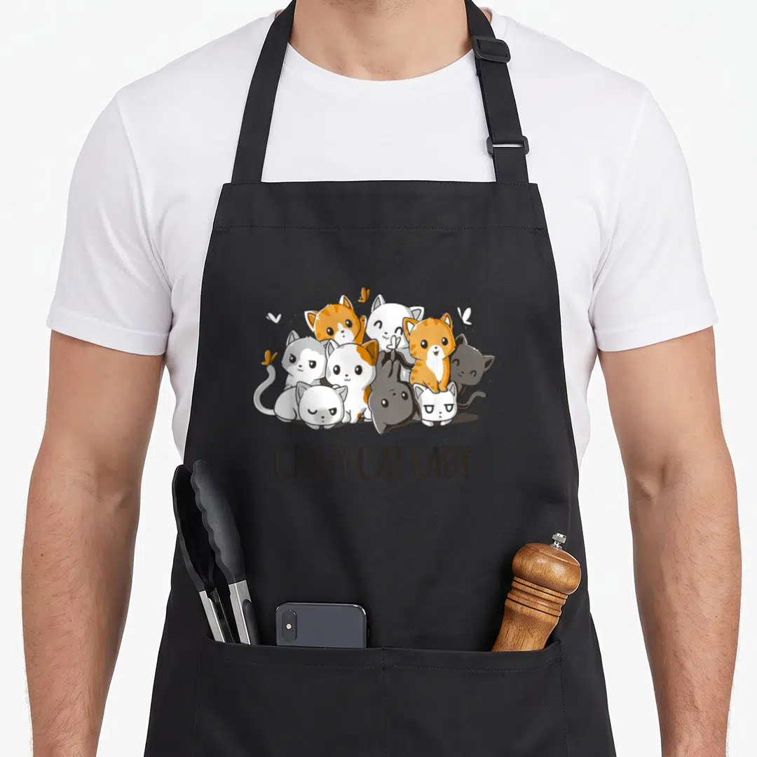 Anime crazy cat lady apron - Ten cartoon cats in orange and grey clustered above the text CRAZY CAT LADY — shown on a black cotton canvas apron against a white background