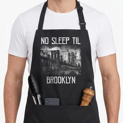 Sleep brooklyn apron - White distressed NO SLEEP TIL BROOKLYN text framing a bridge and skyline graphic — shown on a black cotton canvas apron against a white background