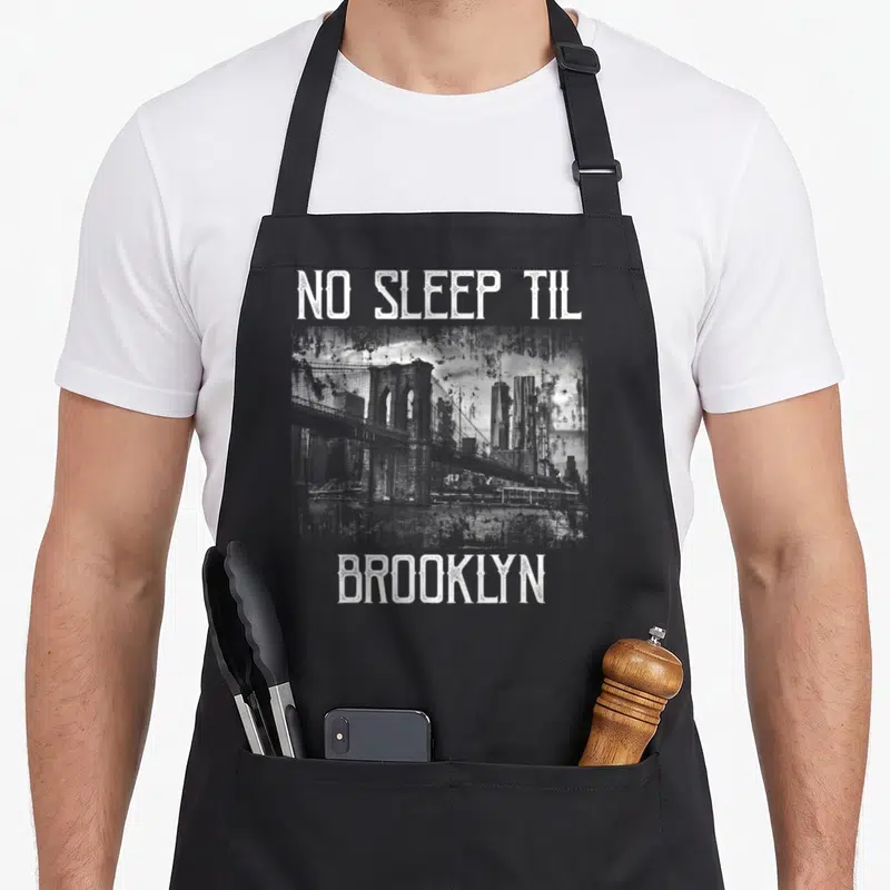 Sleep brooklyn apron - White distressed NO SLEEP TIL BROOKLYN text framing a bridge and skyline graphic — shown on a black cotton canvas apron against a white background