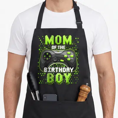 Vintage video birthday apron - Pixelated MOM OF THE BIRTHDAY BOY text surrounding a black game controller on a lime — shown on a black cotton canvas apron against a white background