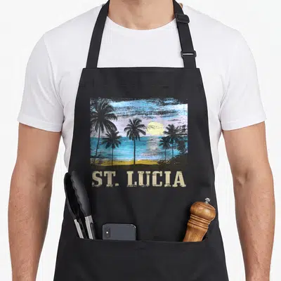 Saint tree apron - Distressed beach scene with palm trees, sun, ocean, and ST. LUCIA text in blue yellow grey beige — shown on a black cotton canvas apron against a white background