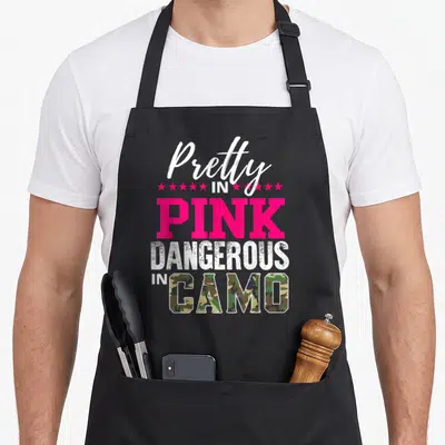Pretty pink camo apron - Stacked text reading Pretty in Pink Dangerous in Camo with pink stars and camo filled letters — shown on a black cotton canvas apron against a white background