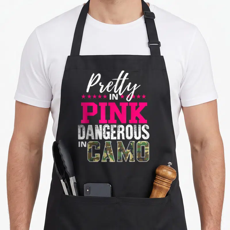 Pretty pink camo apron - Stacked text reading Pretty in Pink Dangerous in Camo with pink stars and camo filled letters — shown on a black cotton canvas apron against a white background