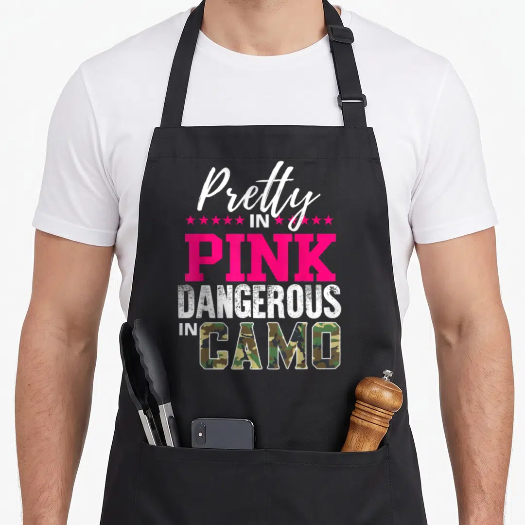 Pretty pink camo apron - Stacked text reading Pretty in Pink Dangerous in Camo with pink stars and camo filled letters — shown on a black cotton canvas apron against a white background