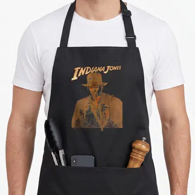 Tonal raiders apron - Indiana Jones Raiders of the Lost Ark realistic portrait with action vignettes in gold and brown — shown on a black cotton canvas apron against a white background