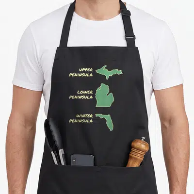 Snowbird winter apron - Three green map outlines labeled Upper, Lower, and Winter Peninsula with yellow text — shown on a black cotton canvas apron against a white background