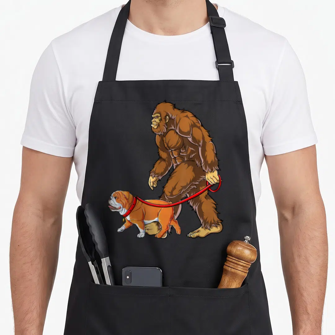 Bigfoot english chef aprons - Brown Bigfoot walking tan bulldog on red leash cartoon — shown on a black cotton canvas apron against a white background