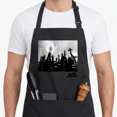 Hand black sabbath apron - Grayscale band photo with raised hands and cross, black text below — shown on a black cotton canvas apron against a white background