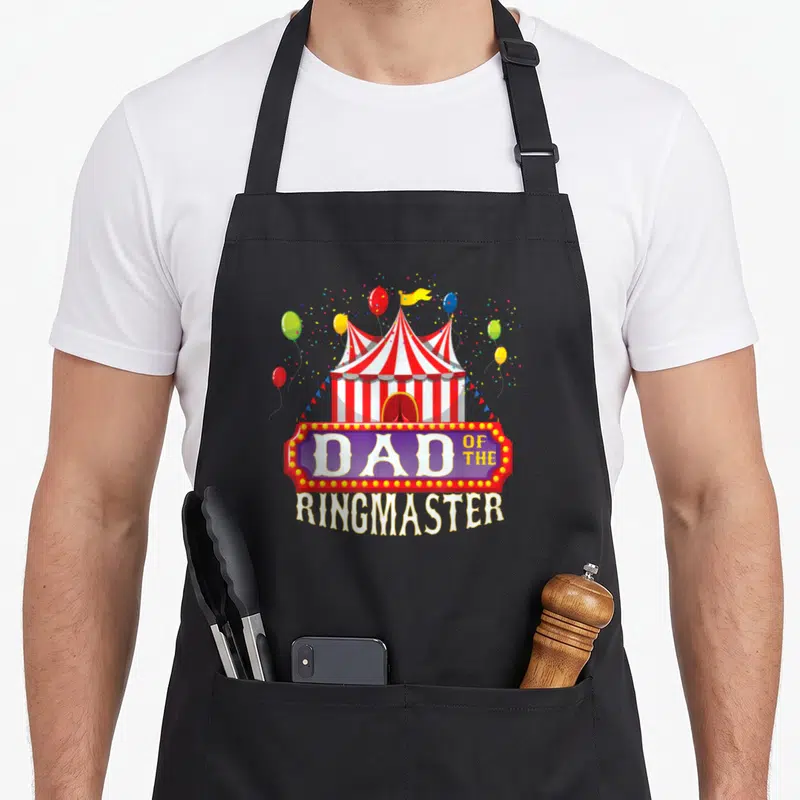 Ringmaster circus birthday apron - Circus tent with balloons and DAD OF THE RINGMASTER marquee sign — shown on a black cotton canvas apron against a white background