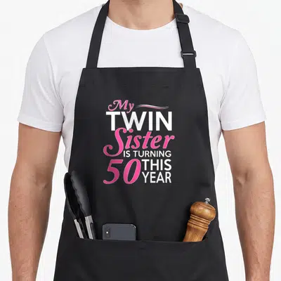 Twin sister birthday apron - Pink and white text reading My Twin Sister Is Turning 50 This Year with decorative swirl — shown on a black cotton canvas apron against a white background