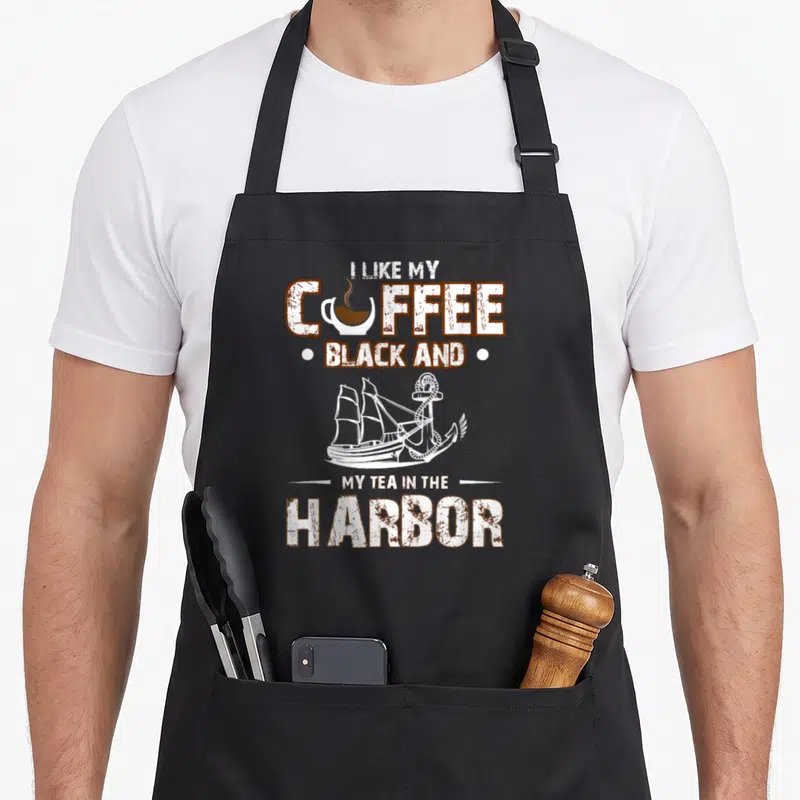 Tea harbor coffee aprons - Distressed brown text reading I like my coffee black and my tea in the harbor with white — shown on a black cotton canvas apron against a white background