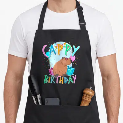 Capybaras birthday apron - Cappy Birthday pun with brown capybara in sunglasses, balloons, and presents — shown on a black cotton canvas apron against a white background