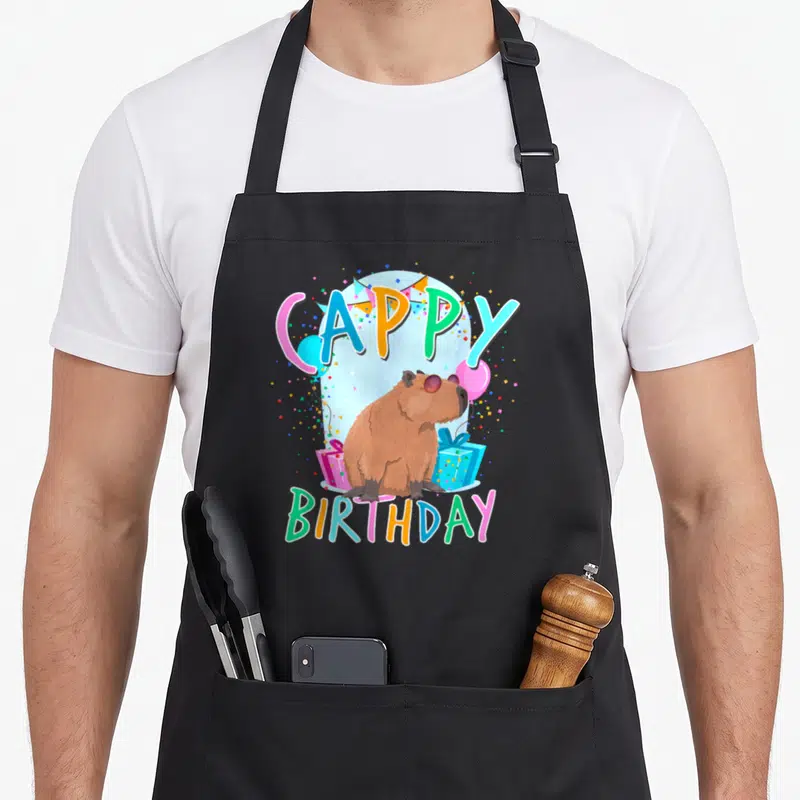 Capybaras birthday apron - Cappy Birthday pun with brown capybara in sunglasses, balloons, and presents — shown on a black cotton canvas apron against a white background