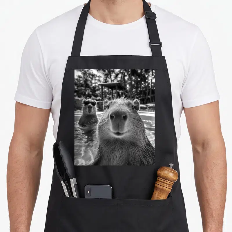 Capybara animal apron - Black and white photo of a capybara face close up with another wearing sunglasses in water — shown on a black cotton canvas apron against a white background