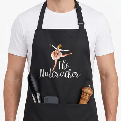 Play nutcracker apron - Peach and white cartoon ballerina posed above stacked white text reading The Nutcracker — shown on a black cotton canvas apron against a white background