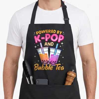Bubble milk aprons - Two bubble tea cups with orange and purple liquid under POWERED BY K-POP AND Bubble Tea text — shown on a black cotton canvas apron against a white background