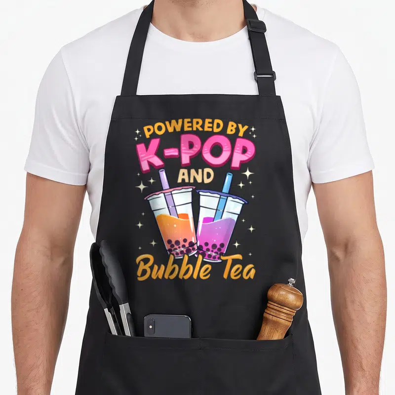 Bubble milk aprons - Two bubble tea cups with orange and purple liquid under POWERED BY K-POP AND Bubble Tea text — shown on a black cotton canvas apron against a white background