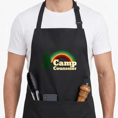 Staff camp chef apron - Rainbow arc next to dark green pine tree with stacked Camp Counselor text — shown on a black cotton canvas apron against a white background