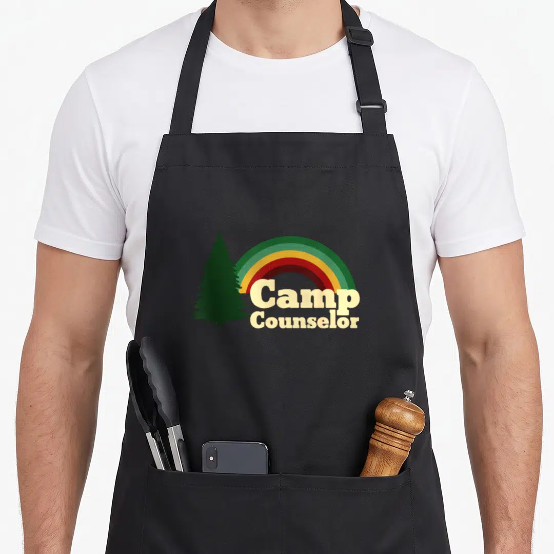Staff camp chef apron - Rainbow arc next to dark green pine tree with stacked Camp Counselor text — shown on a black cotton canvas apron against a white background