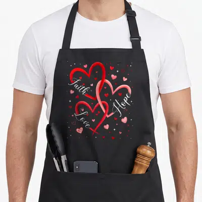 Christian valentine's day apron - Three interlocking red and pink hearts with white Faith Hope Love script — shown on a black cotton canvas apron against a white background