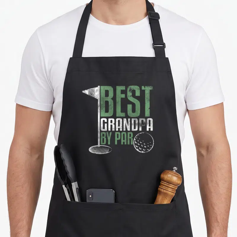 Grandpa grandad aprons - Distressed green and white text best grandpa by par with golf flag hole and ball graphic — shown on a black cotton canvas apron against a white background