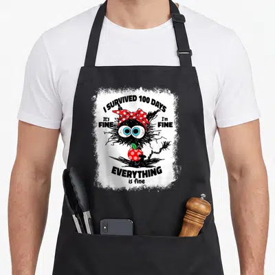 Bleached cat apron - Black monster with red bow holding apple surrounded by sarcastic survival text — shown on a black cotton canvas apron against a white background