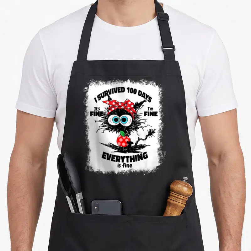 Bleached cat apron - Black monster with red bow holding apple surrounded by sarcastic survival text — shown on a black cotton canvas apron against a white background