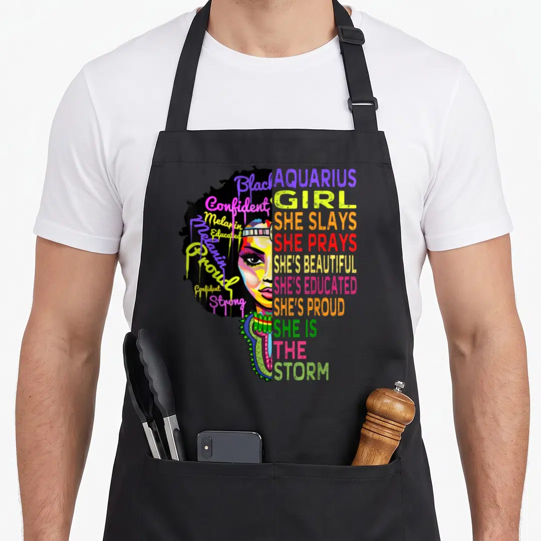 Pray birthday apron - Split face illustration with colorful text reading She Slay She Prays She Is The Storm Aquarius — shown on a black cotton canvas apron against a white background