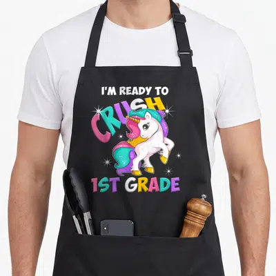 Grade unicorn apron - Cartoon unicorn with multicolored mane and text I'm Ready to Crush 1st Grade — shown on a black cotton canvas apron against a white background
