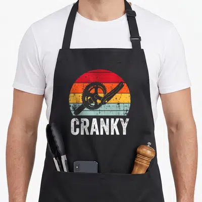 Cranky cycling apron - Retro bicycle crankset over red orange yellow blue striped sun with white text — shown on a black cotton canvas apron against a white background