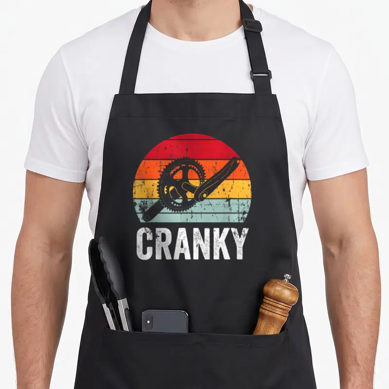 Cranky cycling apron - Retro bicycle crankset over red orange yellow blue striped sun with white text — shown on a black cotton canvas apron against a white background
