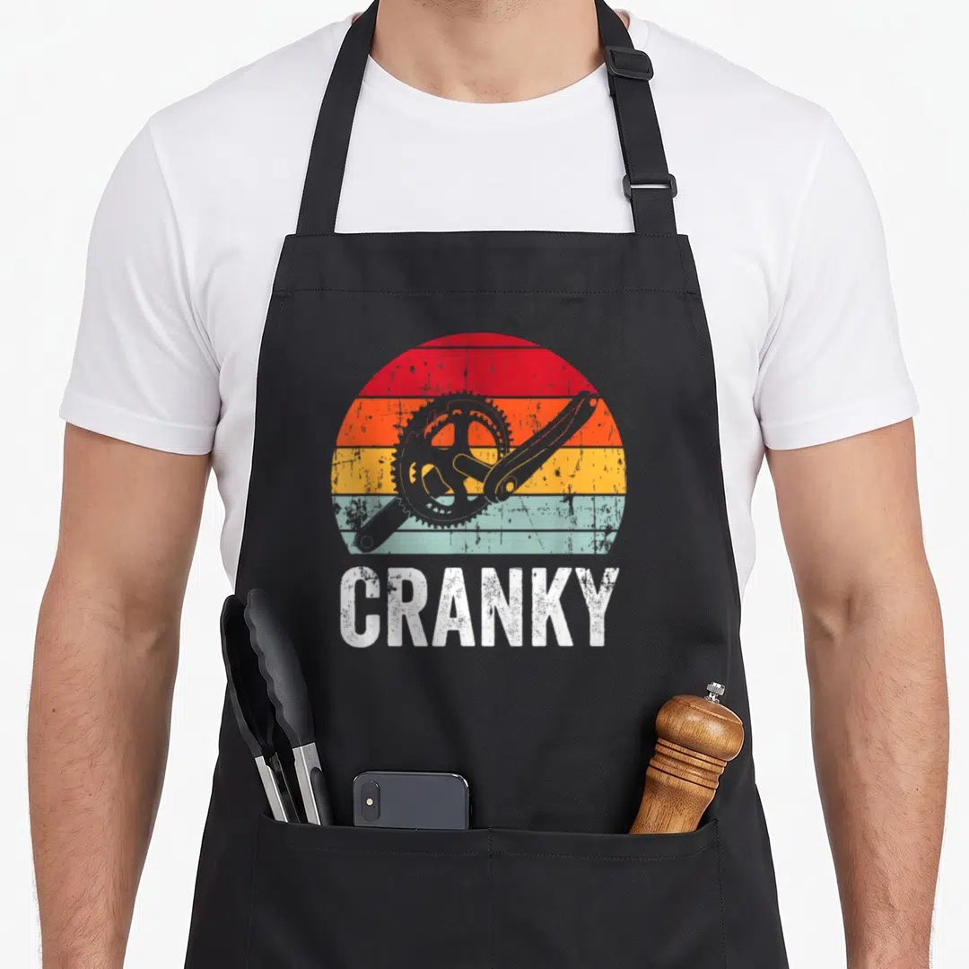 Cranky cycling apron - Retro bicycle crankset over red orange yellow blue striped sun with white text — shown on a black cotton canvas apron against a white background
